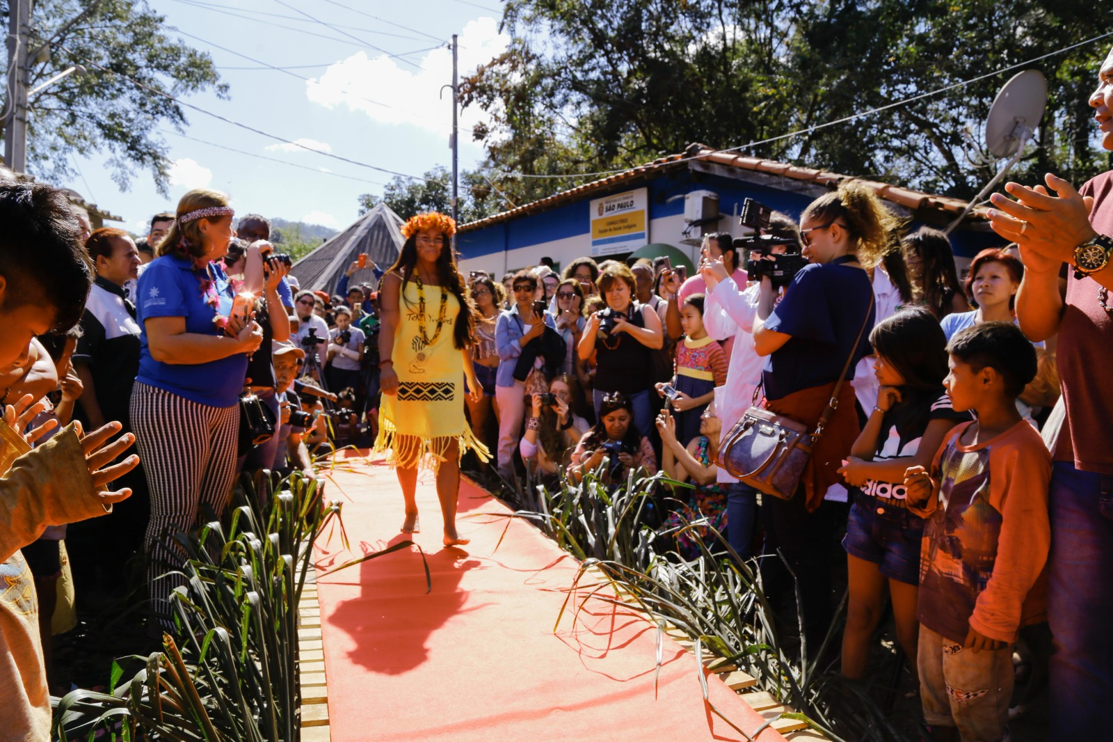 A foto mostra desfile realizado no Dia da Beleza Indígena; na passarela está uma mulher indígena usando cocar e um vestido amarelo